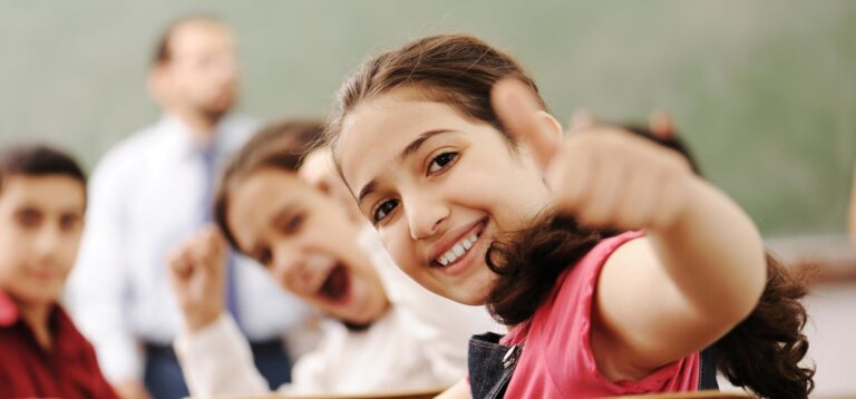 Girl in class rising thumbs up with teacher and classmates in background